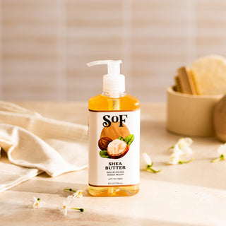 A clear pump bottle of Shea Butter Nourishing Hand Wash stands on a light surface with white flowers and a towel. The BPA free package is softly lit, while a blurred container holding brushes appears in the background.