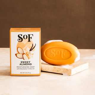 A box of Sweet Almond Moisturizing Bar Soap sits beside an oval bar on a beige surface with a brown background.