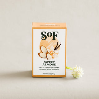 A box of Sweet Almond Moisturizing Bar Soap with organic shea butter and olive oil stands upright on a light surface. A white flower lies beside it, and the box shows images of almonds and a flower on the label.