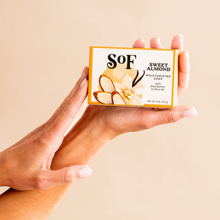 A person holds a box of Sweet Almond Moisturizing Bar Soap featuring almond and flower illustrations on the label, set against a plain beige background.