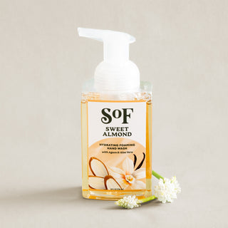 A bottle of Sweet Almond Hydrating Foaming Hand Wash with a white pump rests on a beige surface next to a small white flower. The label shows almond and vanilla illustrations and highlights its paraben-free formula.
