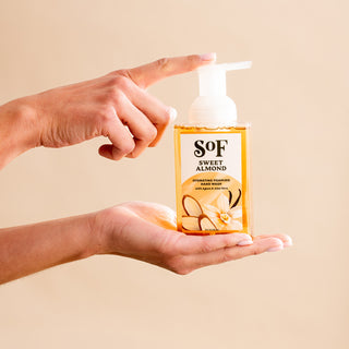 A hand holds a bottle of Sweet Almond Hydrating Foaming Hand Wash as another presses the pump. The clear, SLS/SLES-free bottle displays almond and flower images on its beige label.