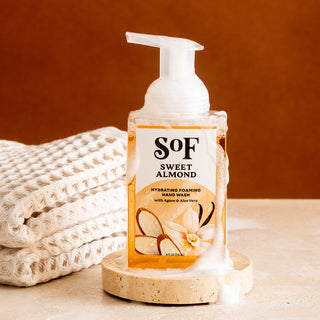 A bottle of Sweet Almond Hydrating Foaming Hand Wash, phthalate and paraben free, sits on a round stone coaster surrounded by foam. Soft cream towels are set behind it against a warm brown backdrop.