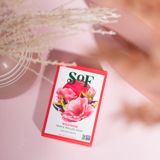 A box of Wild Rose Moisturizing Bar Soap with organic shea butter, decorated with a large pink rose illustration, rests on a soft pink surface beside dried flowers for a nourishing lather experience.