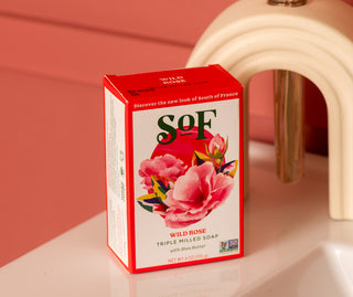 A box of Wild Rose Moisturizing Bar Soap with shea butter and natural colorants, paraben-free, sits on a white surface. Its red and white packaging features pink roses set against a pink background.