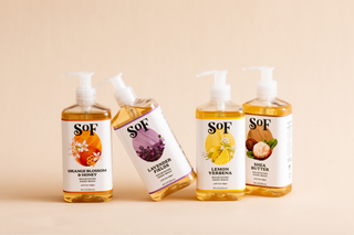 Four SOF brand liquid hand soap bottles with pump dispensers, each featuring a different scent: Orange Blossom & Honey, Lavender Fields, Lemon Verbena, and Shea Butter, set against a beige background.