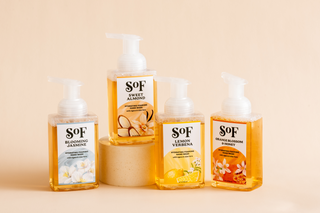 Four SOF brand foaming hand soaps in clear bottles with pump tops, each labeled with different scents: Blooming Jasmine, Sweet Almond, Lemon Verbena, and Orange Blossom & Honey, arranged on a beige surface.