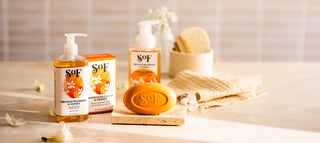 A bottle of liquid soap, a bar of soap, and a box labeled SOF Orange Blossom & Honey sit on a light surface with a textured cloth, a soap dish, and small white flowers in soft, natural lighting.