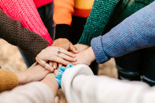 A group of people standing in a circle place their hands together in the center, showing teamwork and unity. Each person is wearing a colorful sweater.